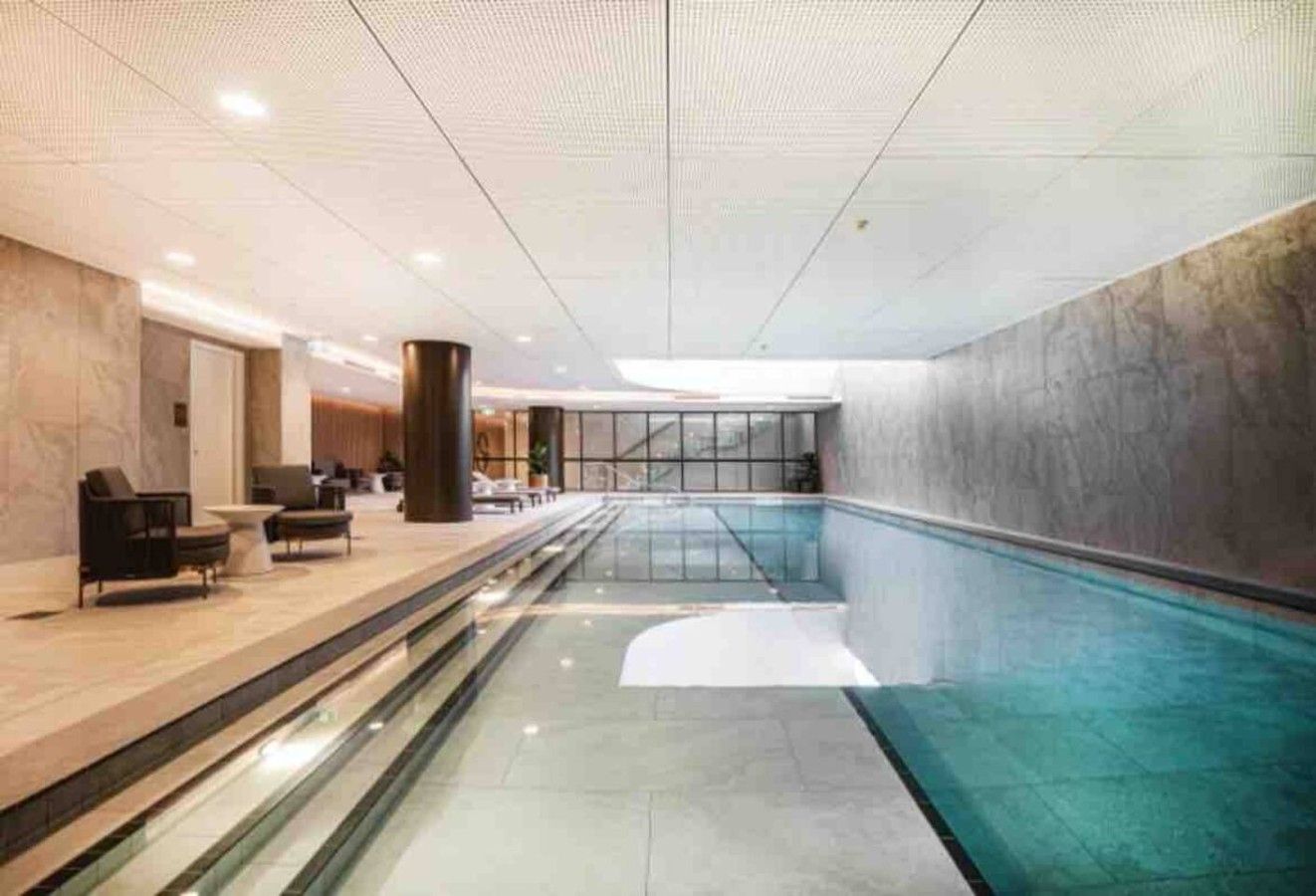 A modern indoor swimming pool with a clear glass barrier, a lounge area with seating, and a high ceiling with recessed lighting.