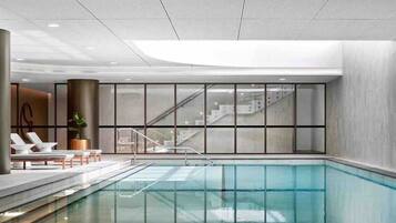 A modern indoor swimming pool with clear glass walls, a staircase leading to upper levels, and lounge chairs along the poolside.