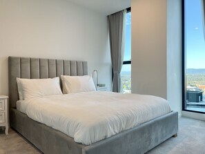Une chambre moderne avec un grand lit, une tête de lit grise et une vue sur le paysage urbain, visible à travers une grande fenêtre.