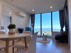 A modern living room with a large window, a dining area with a table and chairs, and a sofa. - SkyGarden Lux Apt | Sunrise View | Pool/Parking (Glen Waverley)