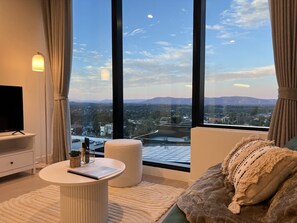 A modern living room with a large window, a flat-screen TV, a round white table, and a comfortable sofa with a textured throw pillow. - SkyGarden Lux Apt | Sunrise View | Pool/Parking (Glen Waverley)