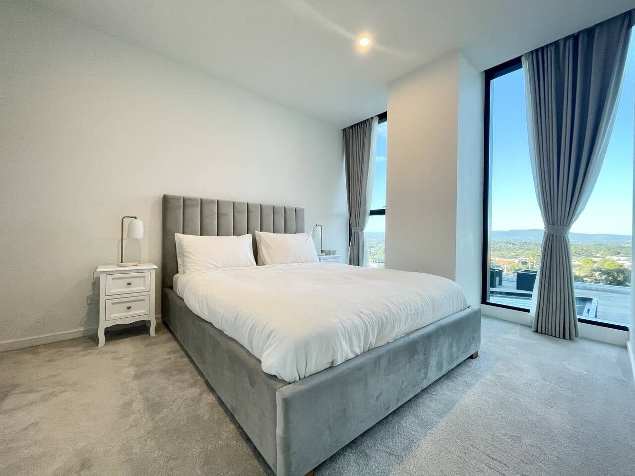 A bedroom with a large bed, a nightstand, a lamp, and a view of the outdoors through a window.