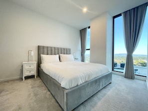 A bedroom with a large bed, a nightstand, a lamp, and a view of the outdoors through a window.
