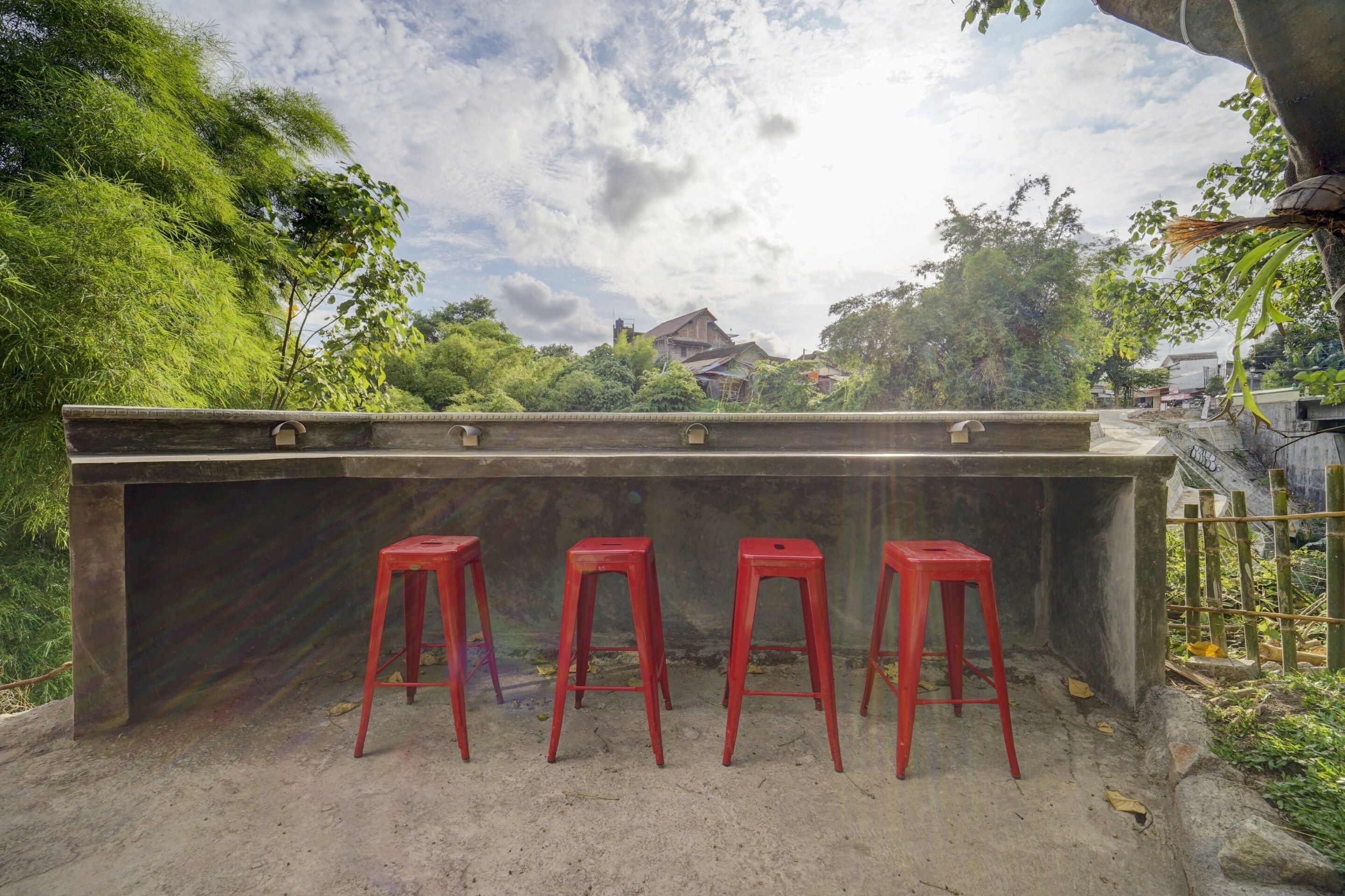 A concrete bench area with four red stools, surrounded by greenery and a cloudy sky.