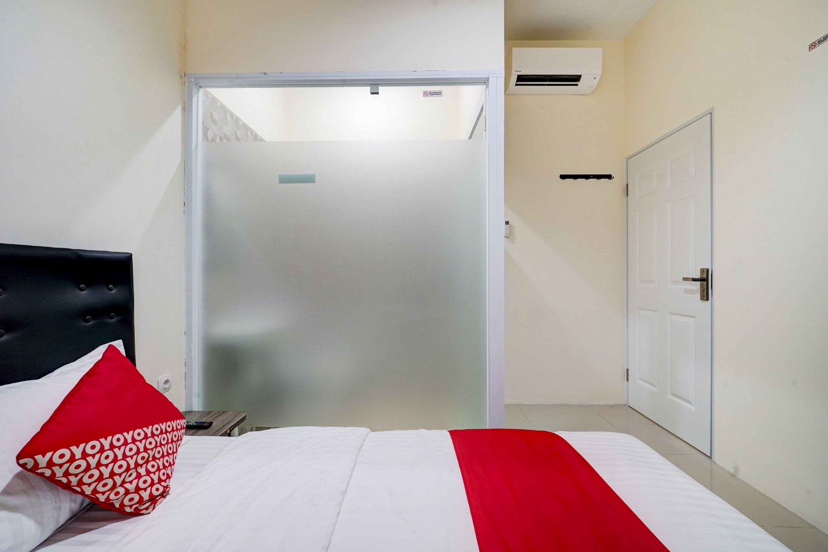 Una habitación de hotel con una cama, una almohada estampada en rojo y blanco, una colcha blanca y roja, una puerta cerrada y un equipo de aire acondicionado en la pared.