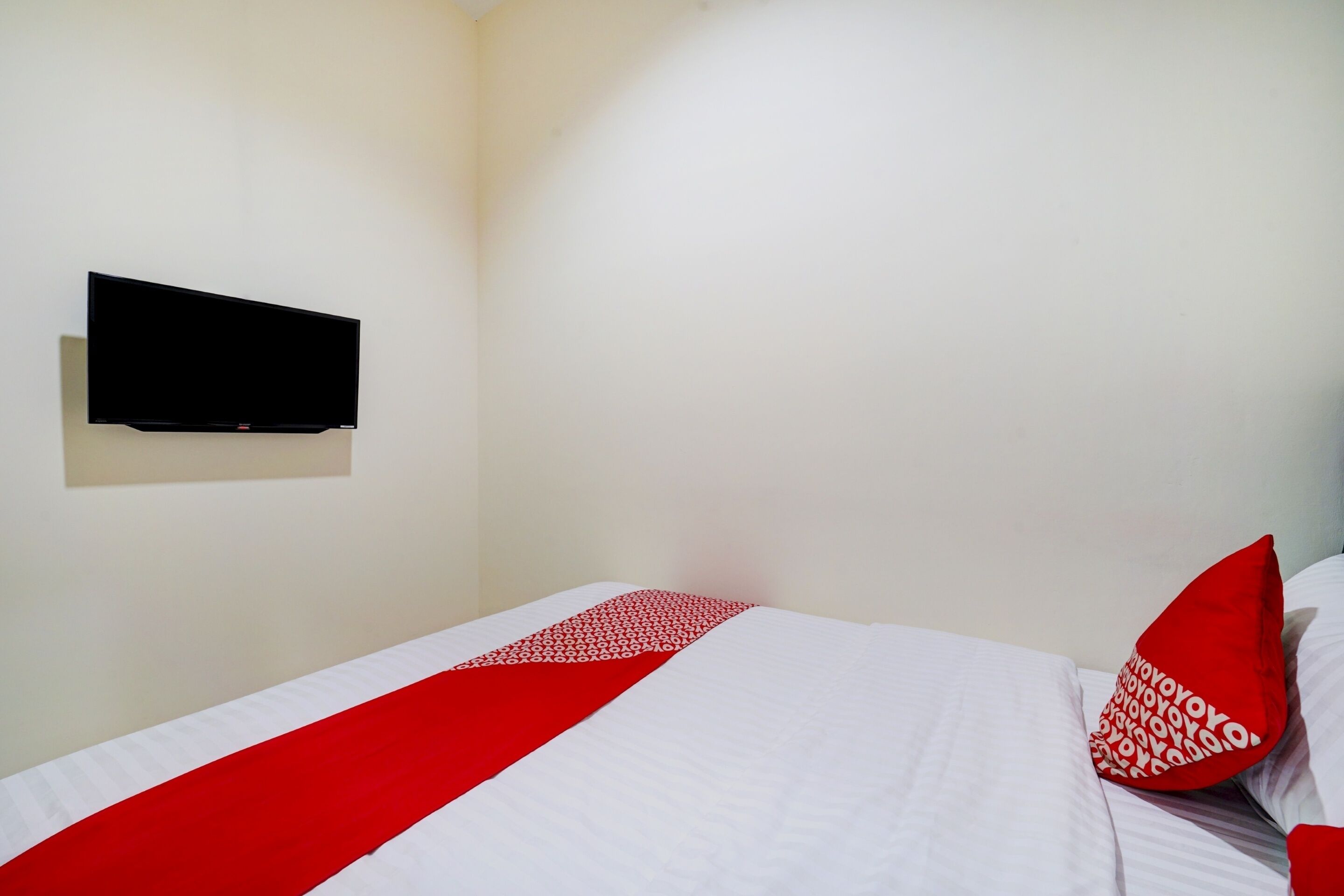 A hotel room with a bed, a flat-screen TV mounted on the wall, and a red and white patterned pillow.