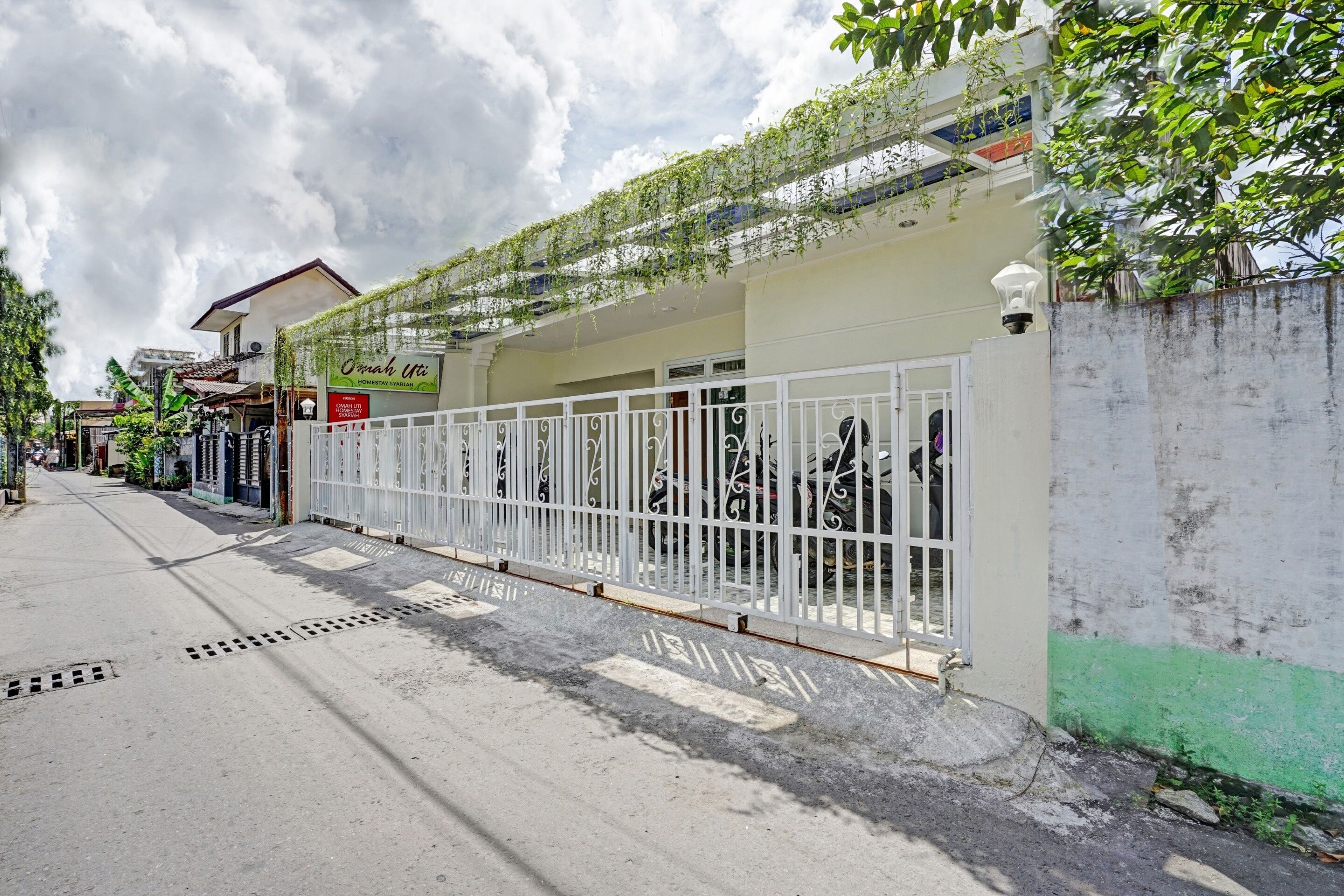 A street view with a gated entrance, a sign for "Omachi Iri," and a building with a green wall and vines.