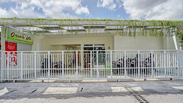 Une entrée fermée par un portail mène à un bâtiment sur lequel est apposée une enseigne indiquant : « Omanh Uti Homestay Syariah ».