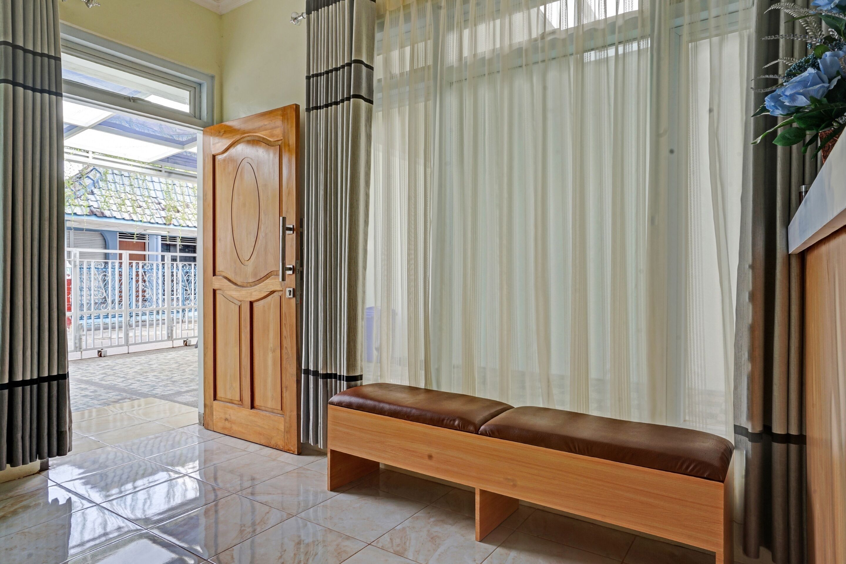 Une pièce avec un banc en bois, une grande fenêtre ornée de rideaux légers et une porte donnant sur l’extérieur.