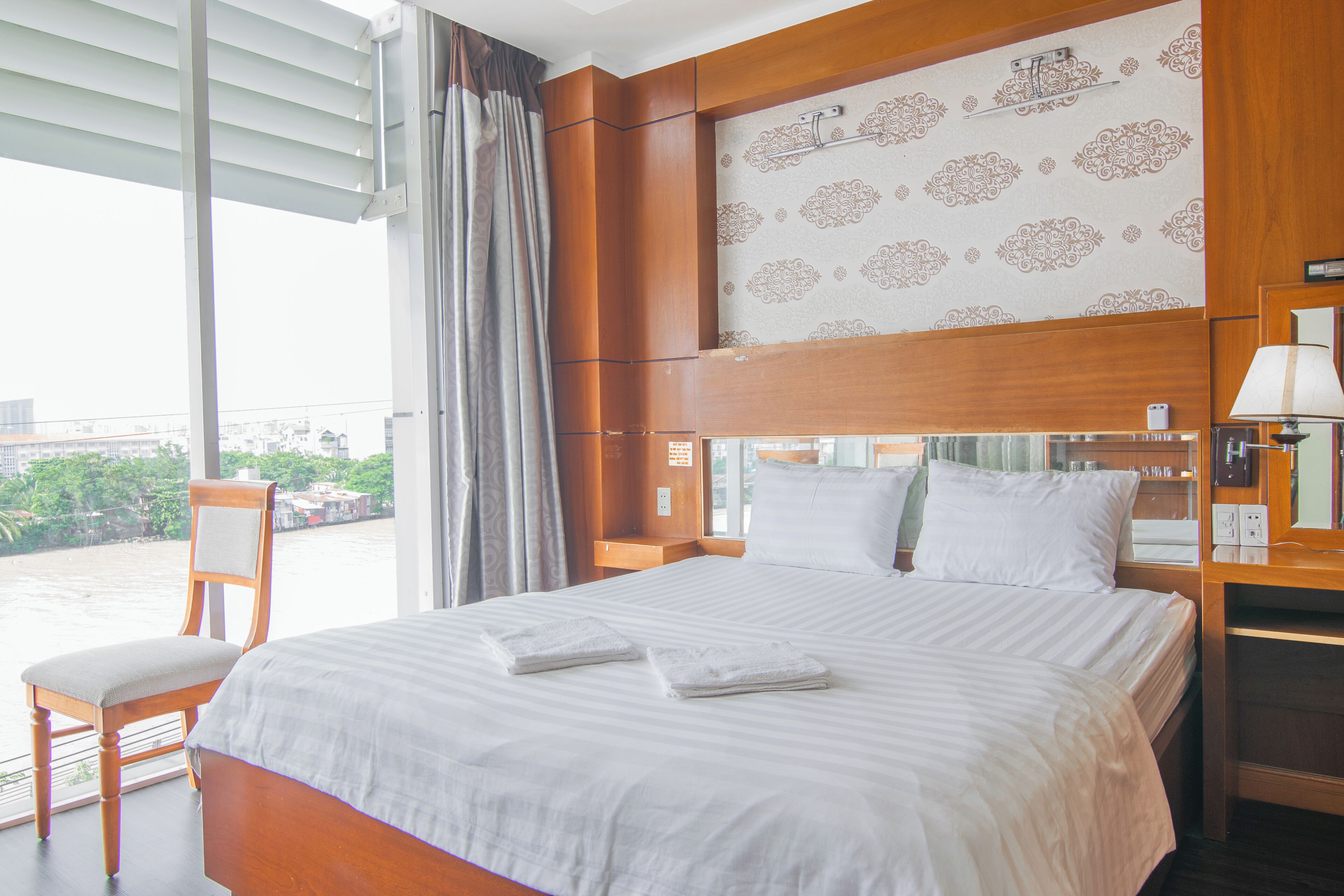 A hotel room with a large bed, a chair, a nightstand, and a view of the city through the window.