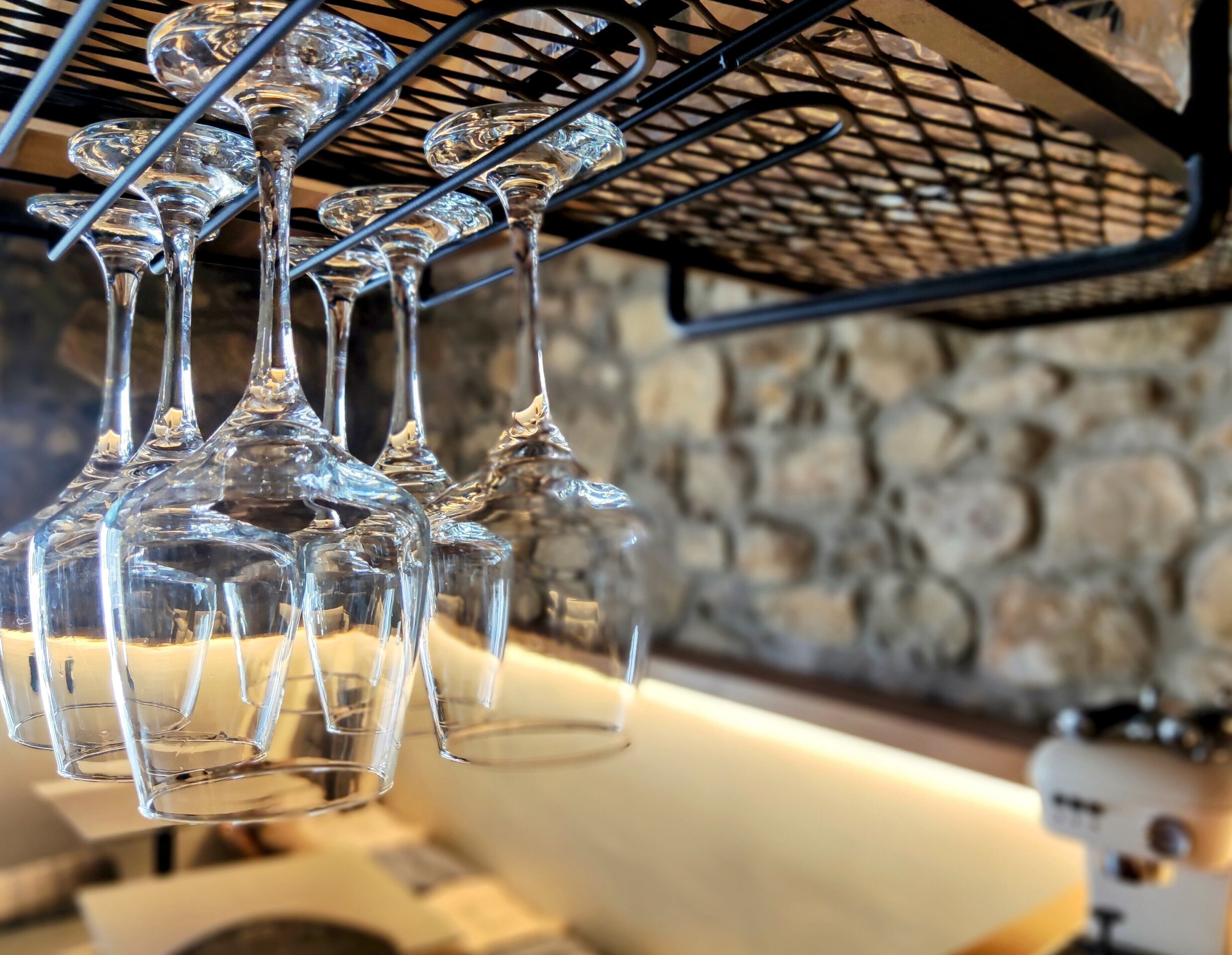A collection of clear wine glasses hanging from a black metal rack against a stone wall.