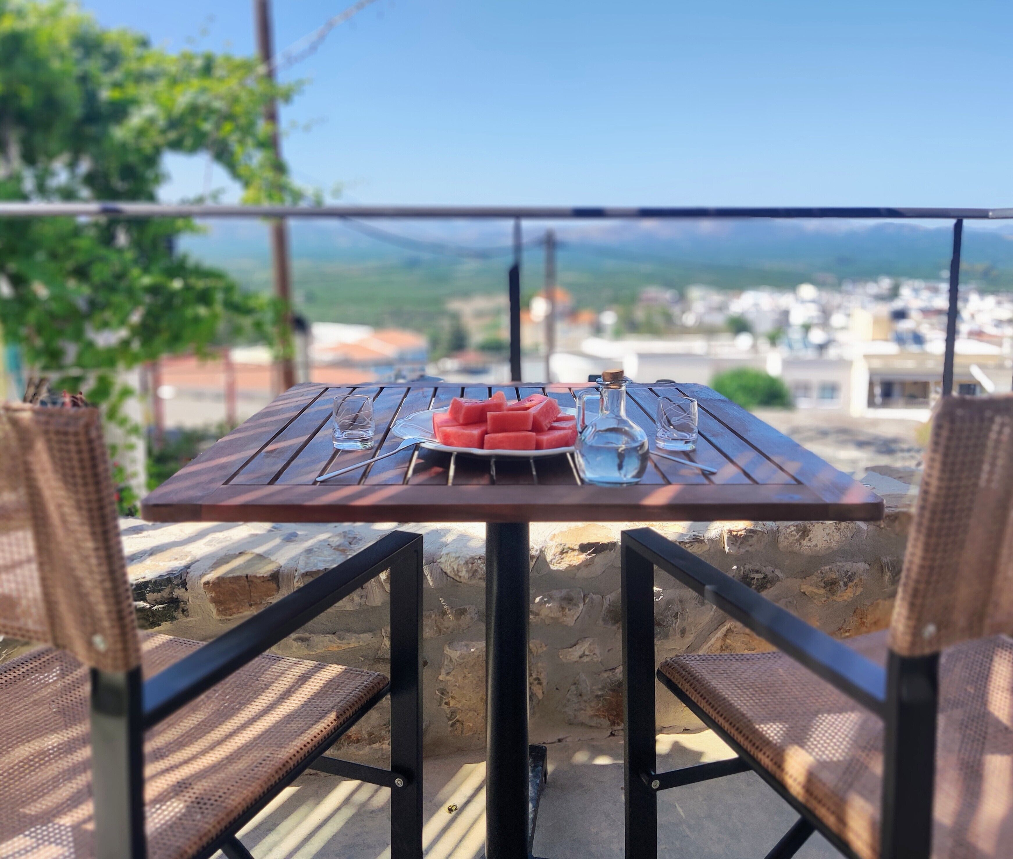 Une table avec une assiette de pastèque, un verre et une bouteille, placée sur une terrasse en plein air d’où l’on peut admirer des bâtiments et des montagnes.