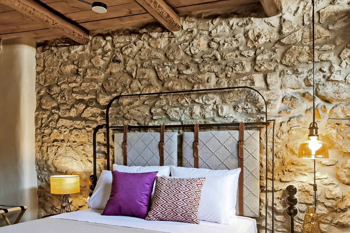 Ein Bett mit Metallgestell, weißer Bettwäsche und mehreren Kissen, darunter ein lilafarbenes. Auf dem Nachttisch steht eine Lampe und neben dem Bett ein dekorativer Flakon. Der Raum zeichnet sich durch eine Steinwand und Holzbalkendecke aus.