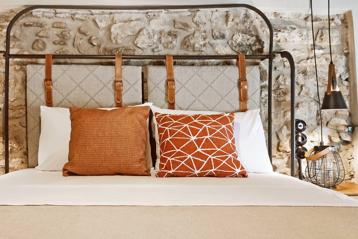Ein Bett mit rustikaler Steinwand, einem Metall-Kopfteil und zwei Kissen.