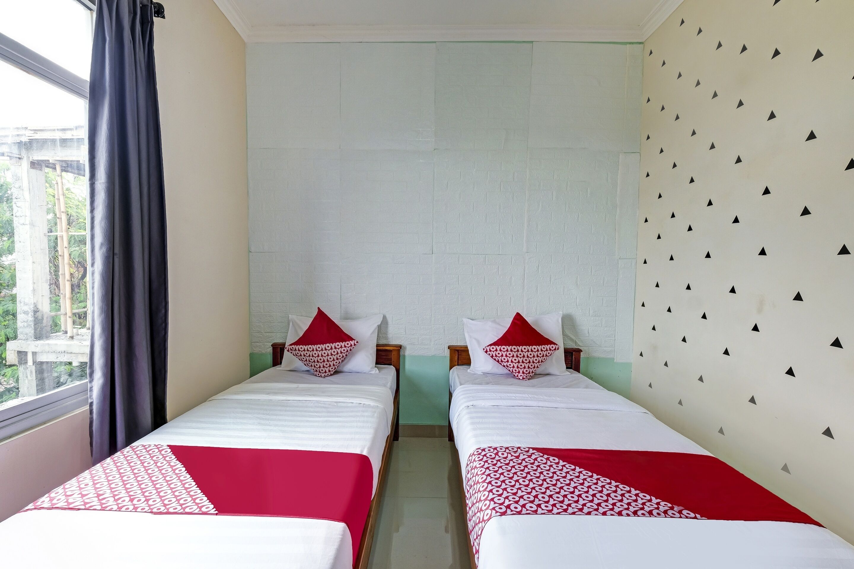 A room with two beds, each with a red and white bedspread and matching pillows. The room has a white brick wall and a window with a curtain.