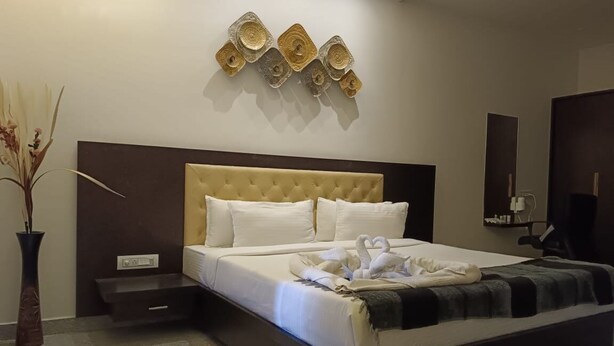 A modern hotel room with a tufted headboard, a large bed with white linens, a decorative wall art piece, a vase with dried plants, and a small bedside table. - Amazing Fun World By Nexottel (Dwarka)