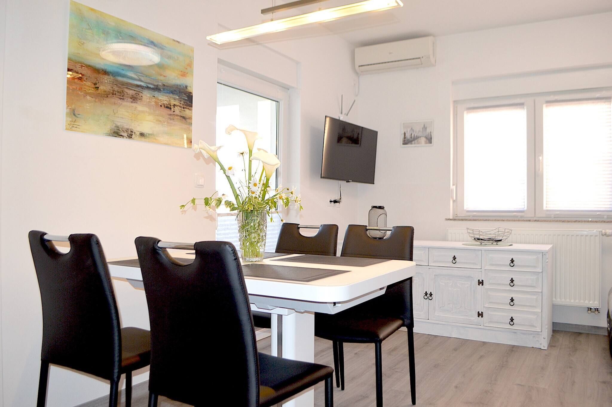 Een eethoek met een witte tafel, zwarte stoelen, een vaas met bloemen, een televisie aan de muur en een raam met zonwering.