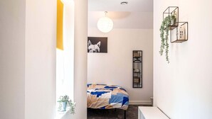 Un couloir étroit avec un lit au fond, une console blanche, un luminaire suspendu et un tableau encadré au mur.