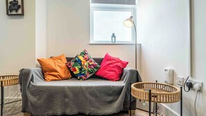 A cozy living area with a grey sofa, colorful pillows, a small table, and a window with a lamp. - 1 bed Apartment near Liverpool Lime street (Old Swan)