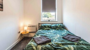 A small, neatly arranged bedroom with a bed, a small table with a lamp, a radiator, and a window with a view of trees and buildings. - 1 bed Apartment near Liverpool Lime street (Old Swan)