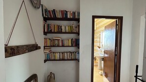 Um corredor estreito com uma estante cheia de livros, uma cadeira rústica e uma porta que dá para um banheiro.