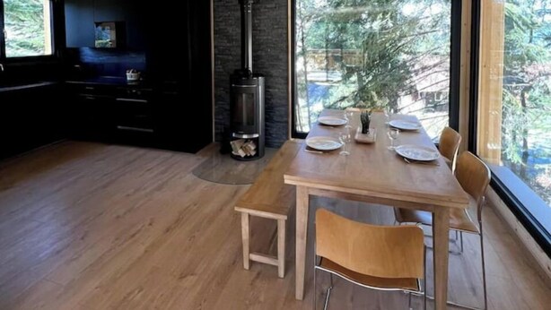 A modern interior with a wooden ceiling, a dining area with a table and chairs, a built-in fireplace, and large windows offering a view of the outdoors.