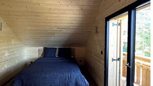 A bedroom with a wooden ceiling, a bed with a dark blue blanket, and a large window with a view of the outdoors.