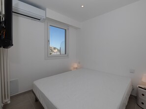 Une chambre à coucher comprenant un lit, une fenêtre, un climatiseur fixé au mur, une table de chevet avec une lampe et un radiateur.