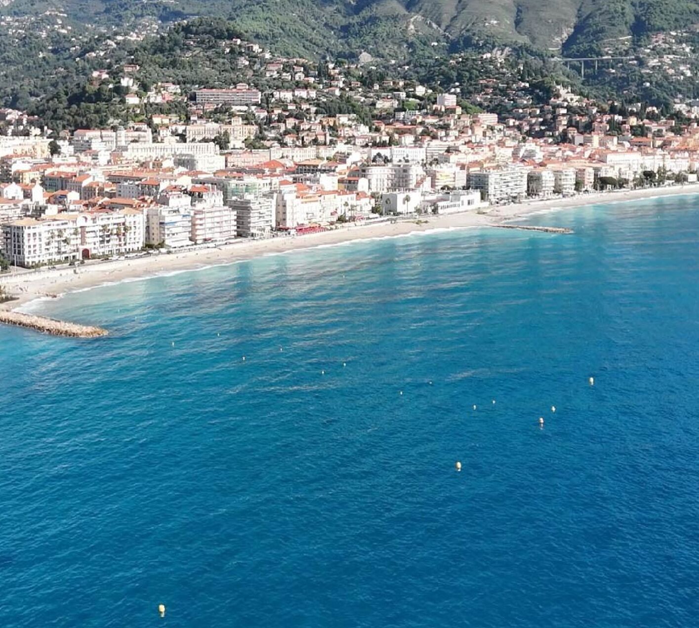 A coastal cityscape with buildings along the waterfront, a clear blue sea, and mountains in the background.