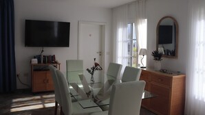 Een moderne woonkamer met een glazen eettafel, witte stoelen, een flatscreen televisie, een boekenkast en een grote spiegel.