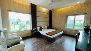 A modern hotel room with a large bed, two bedside tables, a chair, and large windows offering a view of the outdoors. - Hotel Riya Palace (Haldwani)