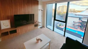 A modern living room with a flat-screen TV, a white coffee table, and a large window offering a view of a swimming pool and buildings. - Modern 1-bedroom Unit in Dicksonnear Shops (Dickson)