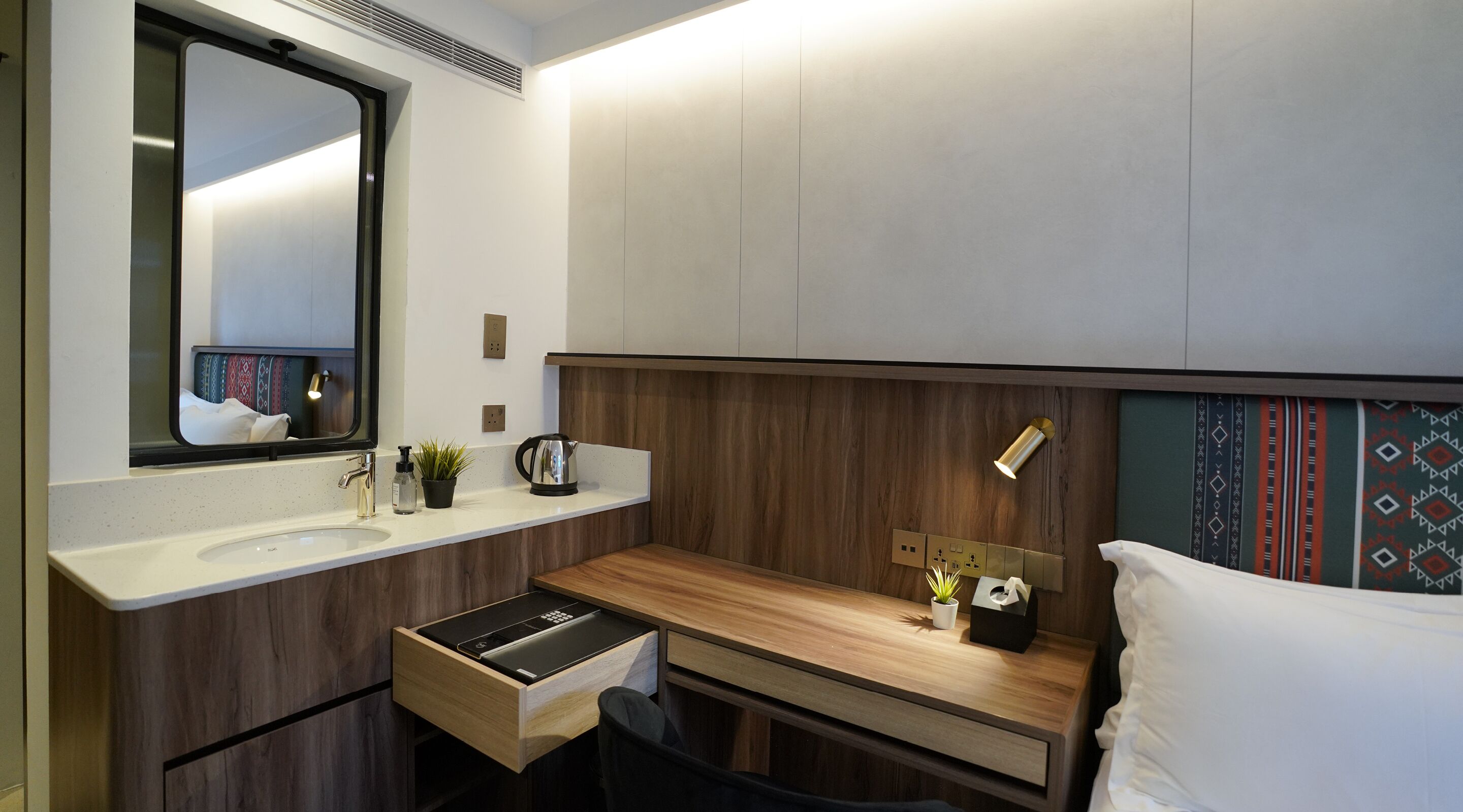 Un baño moderno con tocador de madera, un lavamanos, un espejo, una planta pequeña, una tetera y una mesita de noche con una lámpara.