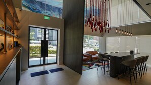 Un moderno vestíbulo de hotel con puerta de vidrio, una zona de estar con mesa y sillas, y un techo con lámparas colgantes.