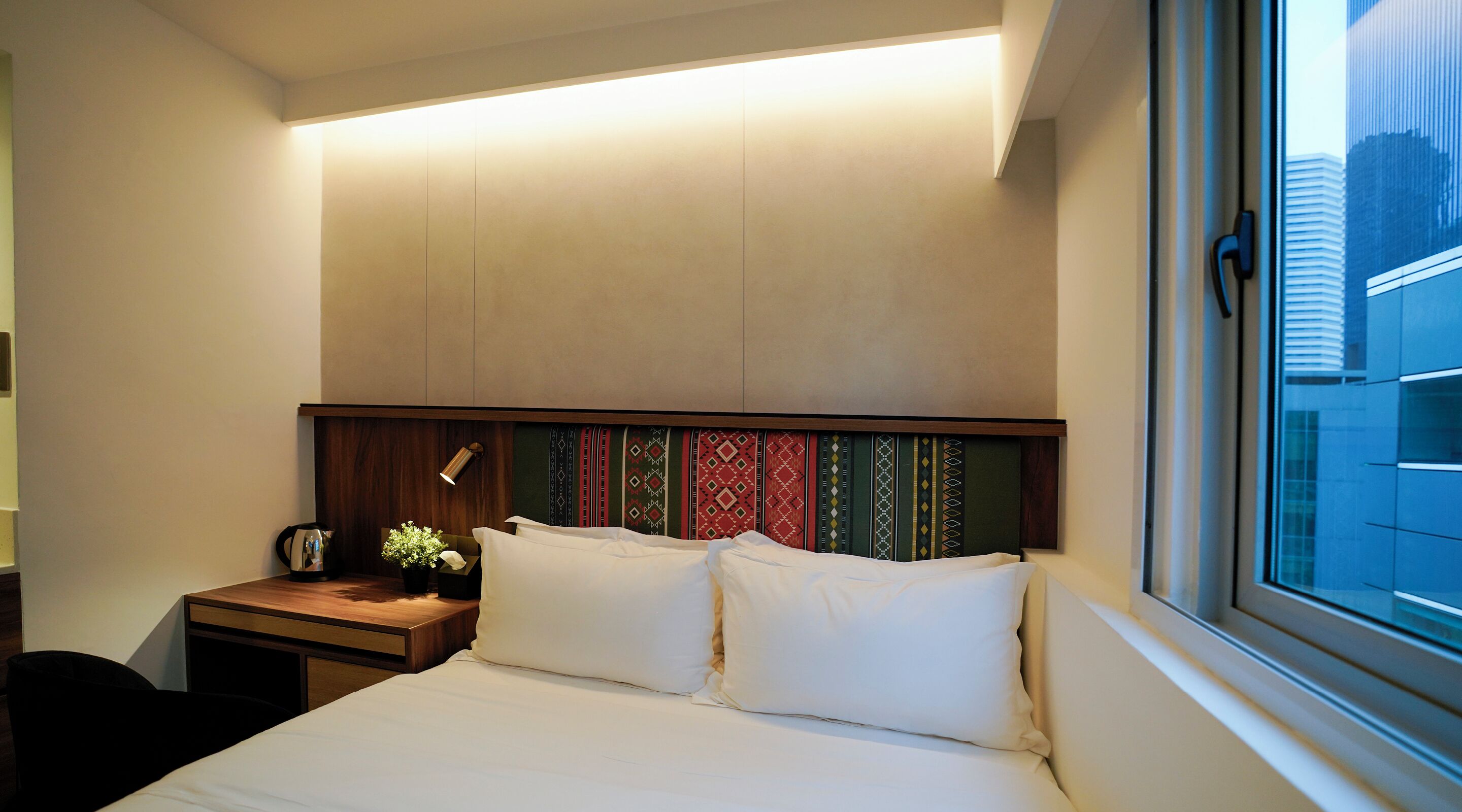 A hotel room with a bed, bedside table, lamp, and a window with a city view.