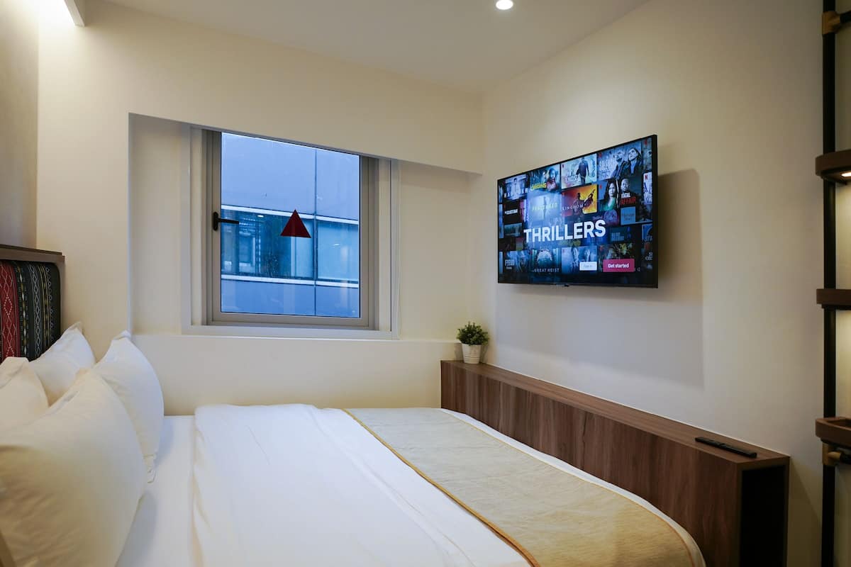 A hotel room with a large bed, a TV mounted on the wall displaying a 'Thrillers' channel, a window with a view of the city, and a small plant on a nightstand.