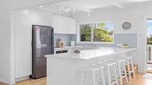 A modern kitchen with white cabinetry, a stainless steel refrigerator, a large window, and a bar area with stools.
