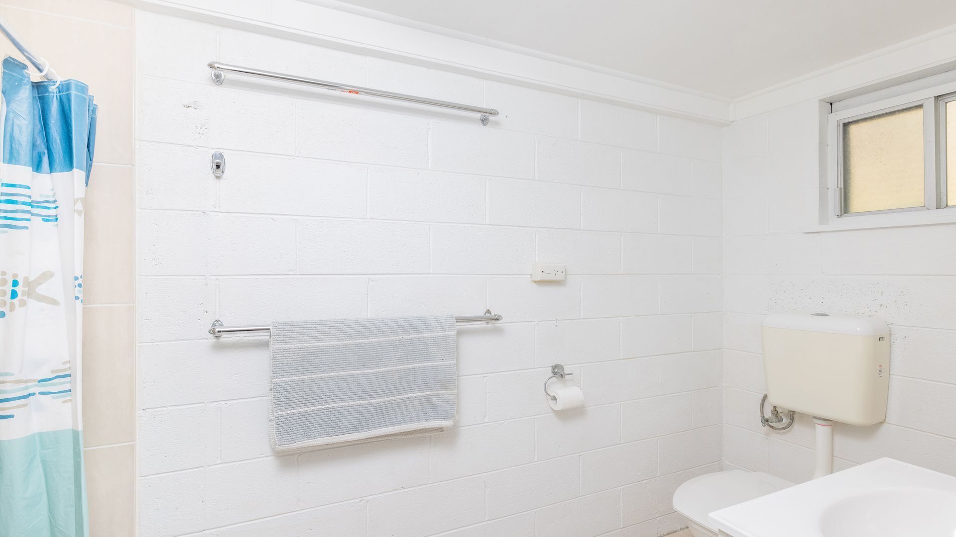 A bathroom with white tiled walls, a white toilet, a towel rack, a shower curtain, and a window.