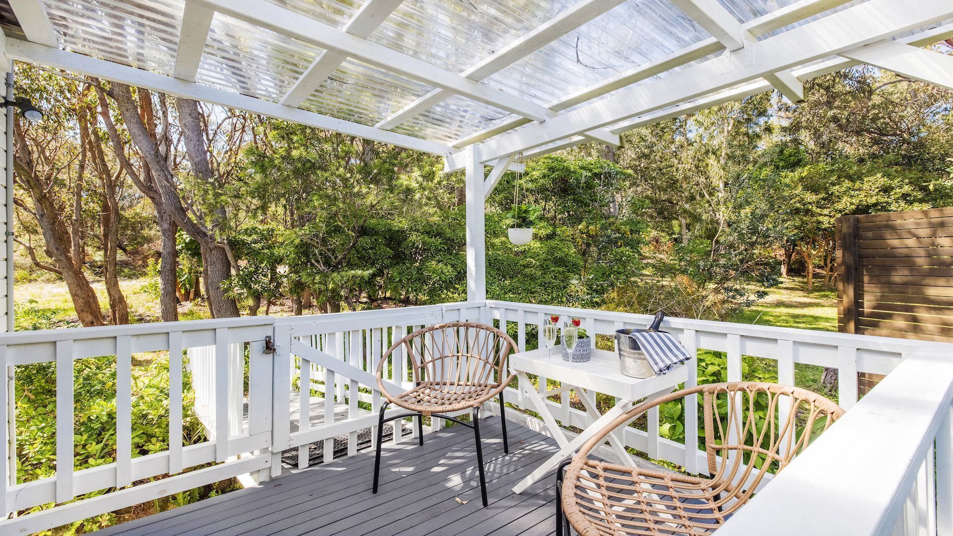 A covered outdoor deck with white railings, two wicker chairs, a small table with a glass, and a hanging planter.