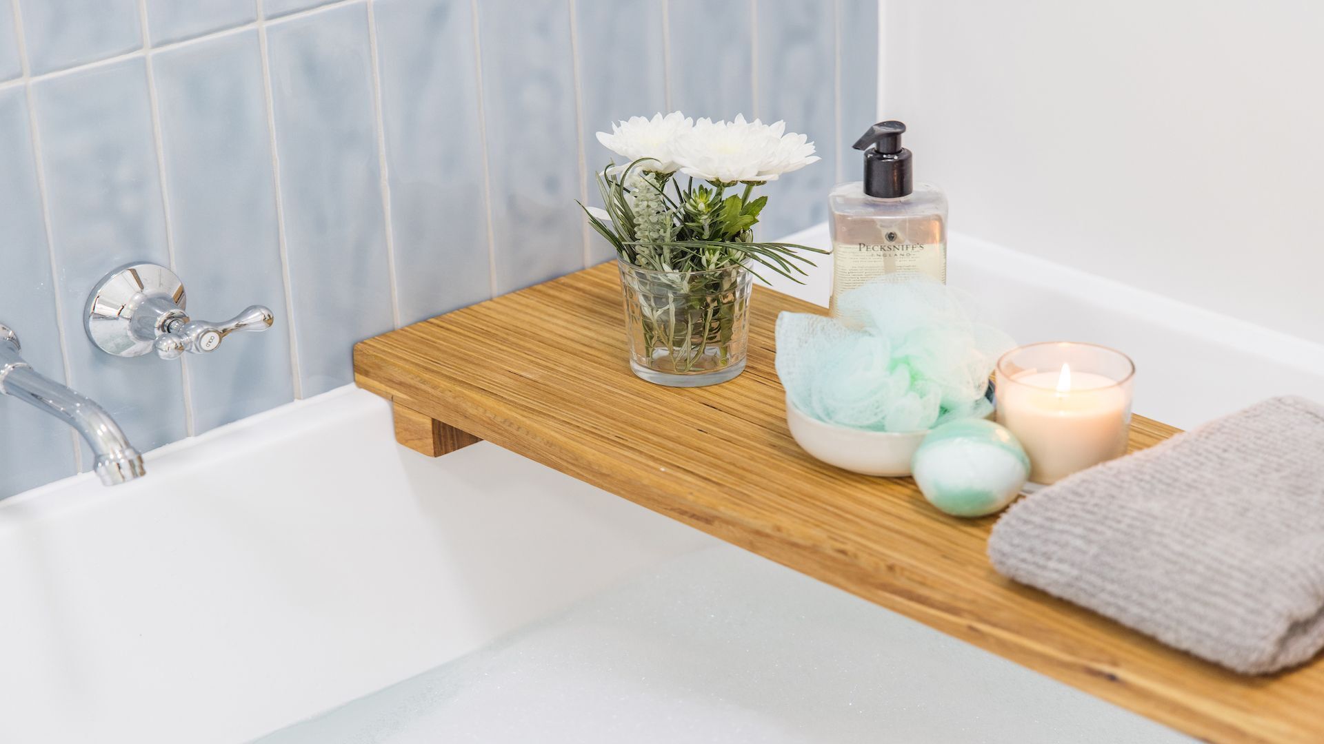A bathroom with a wooden shelf holding a glass vase with flowers, a bottle of hand soap, a candle, and a towel.