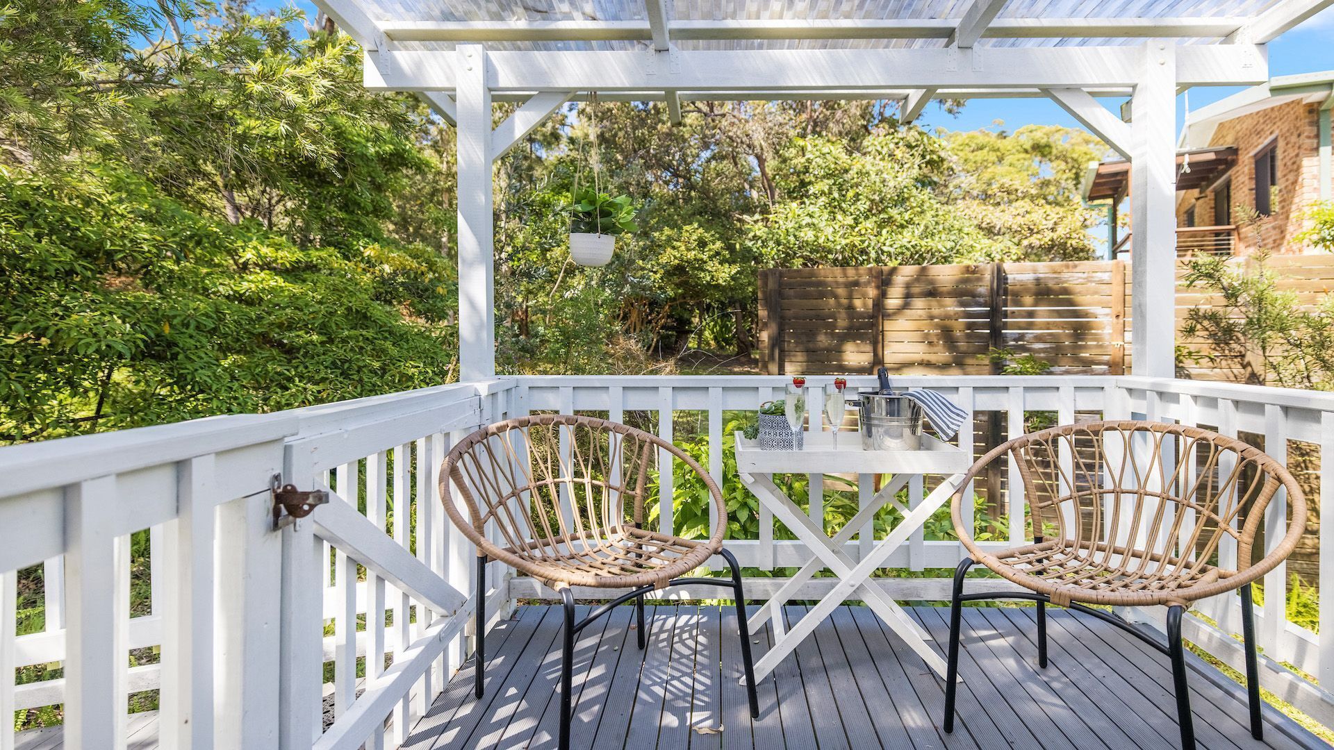 Een overdekt terras buiten met twee rieten stoelen en een tafel, omgeven door witte leuningen en begroeiing.