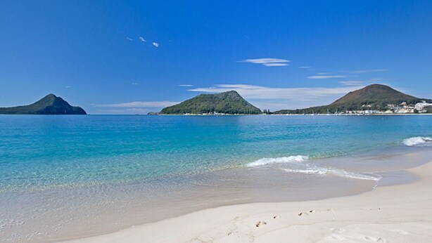 A beach with clear turquoise water, white sand, and two green hills in the background.