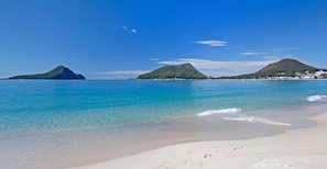 A beach with clear turquoise water, white sand, and two green hills in the background.