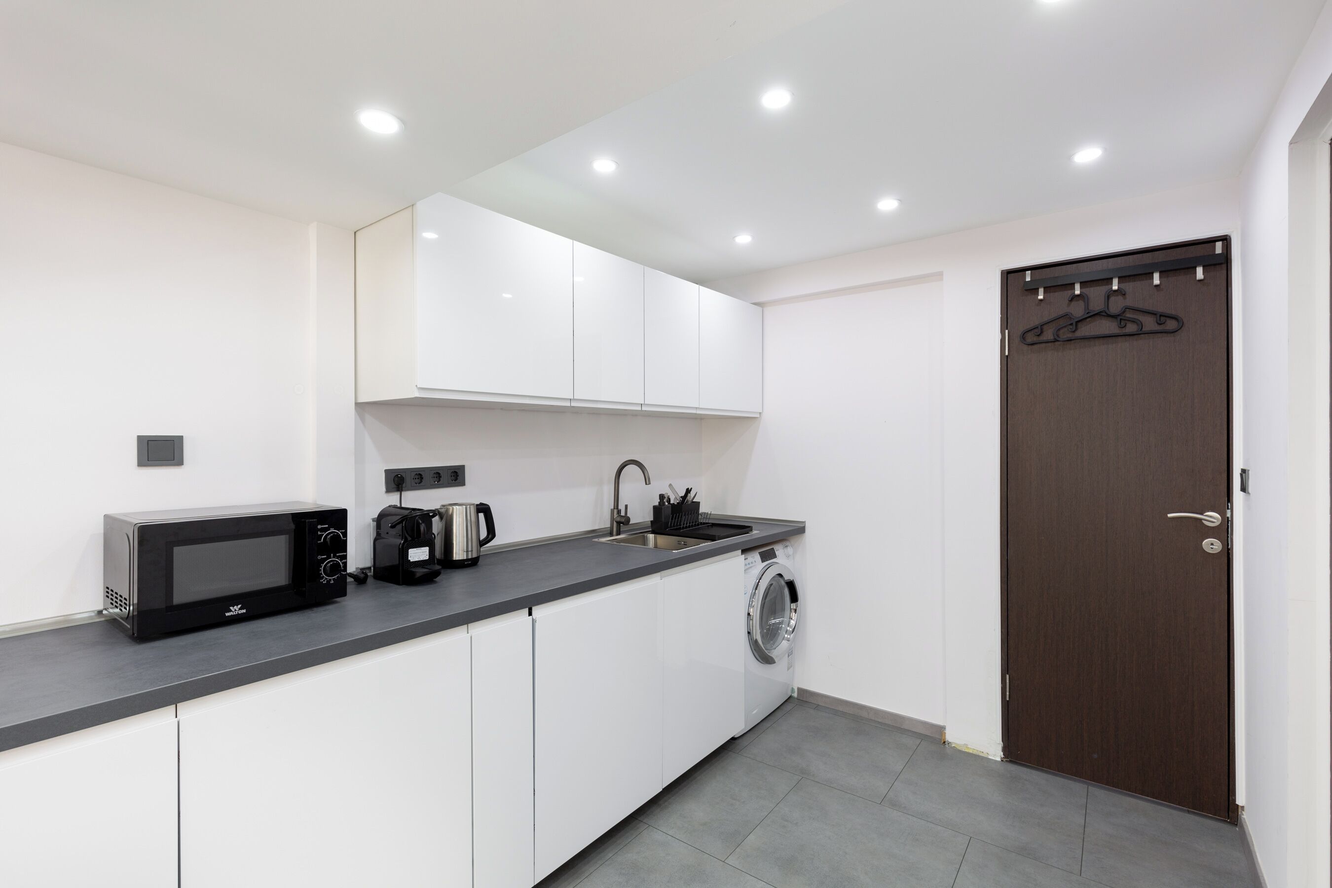 Una cocina compacta con armarios blancos, microondas, cafetera y lavadora.