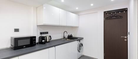 Una cocina compacta con armarios blancos, microondas, cafetera y lavadora.