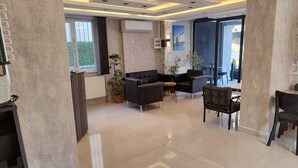 A spacious living area with a black sofa set, a small round table, and a chair. There are large windows providing natural light and a view of greenery outside. - Cam Suites (Kahramanmaras)