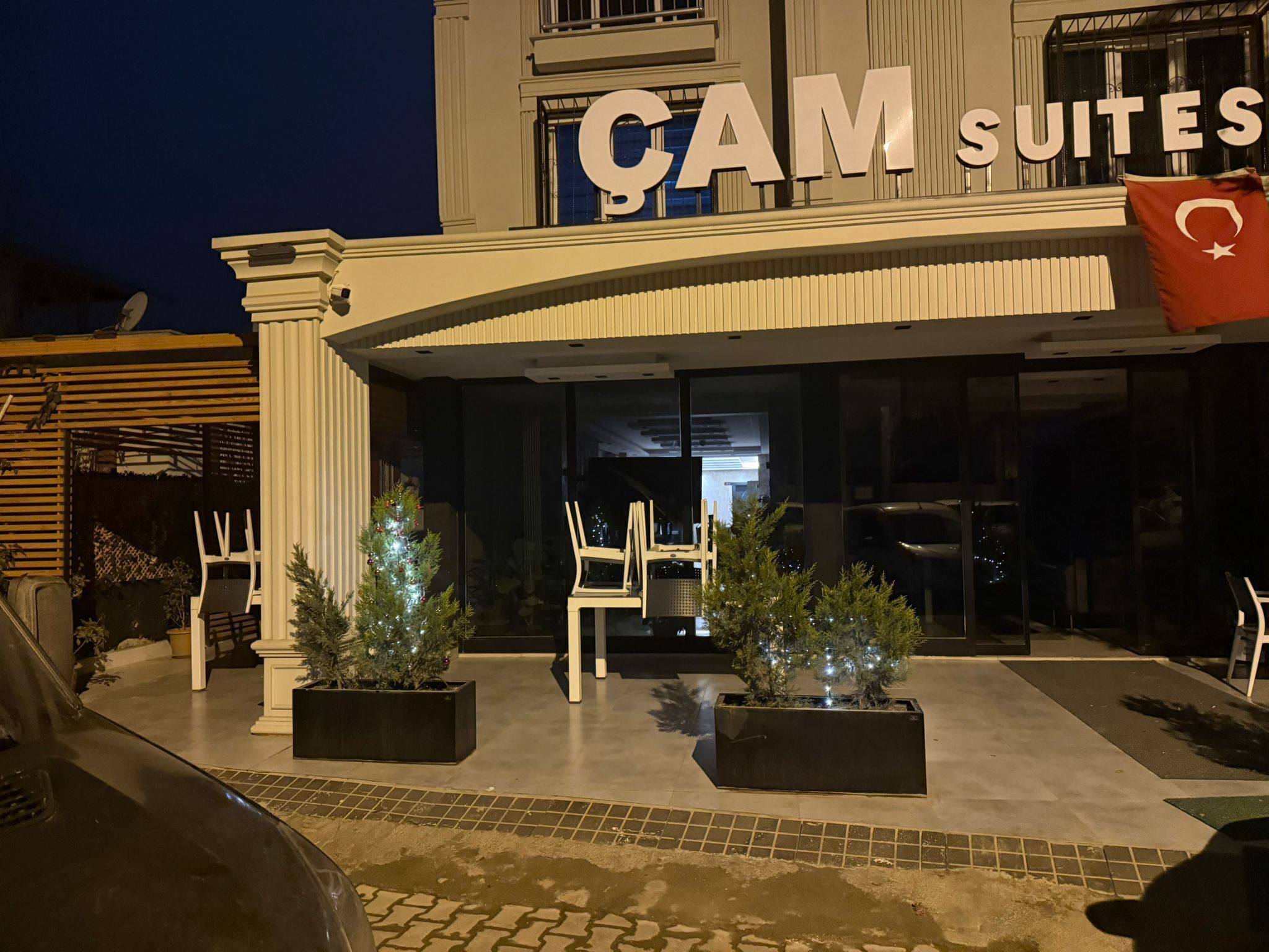 Ein Hotel Eingang mit dem Schild "CAM SUITES" und einer Flagge. Draußen befinden sich Sitzbereiche mit Stühlen und Blumentöpfen.