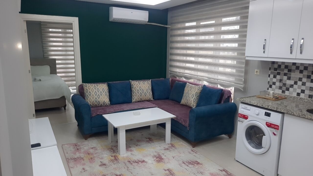 Un ambiente compatto con divano, un piccolo tavolo, lavatrice e angolo cottura.