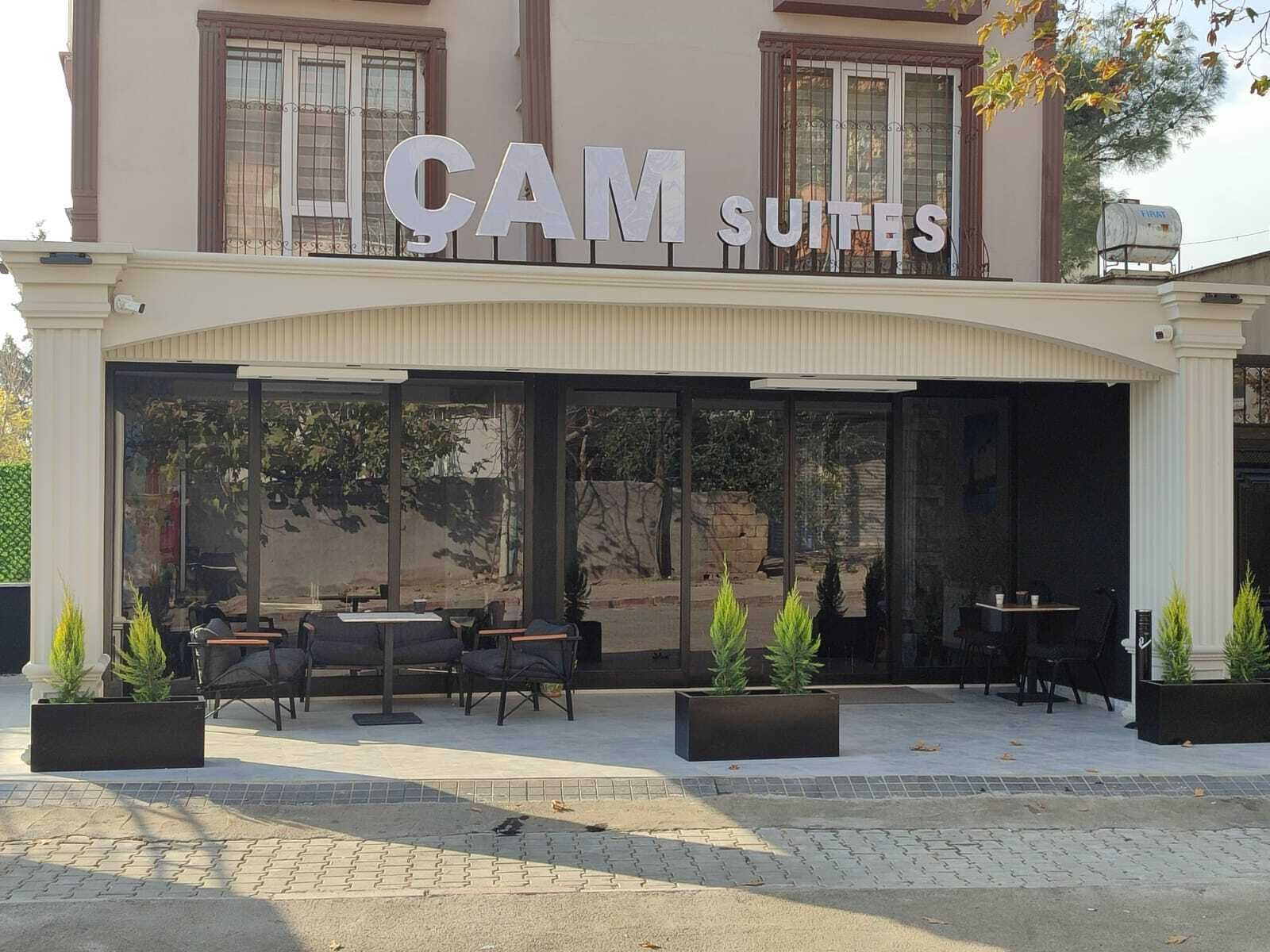Exterior de un hotel con el letrero "CAM SUITES" y mobiliario exterior, incluyendo mesas y sillas.