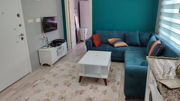 Uma sala de estar com parede verde-água, um sofá branco com almofadas coloridas, uma mesa de centro branca, uma TV em um móvel e uma máquina de lavar.
